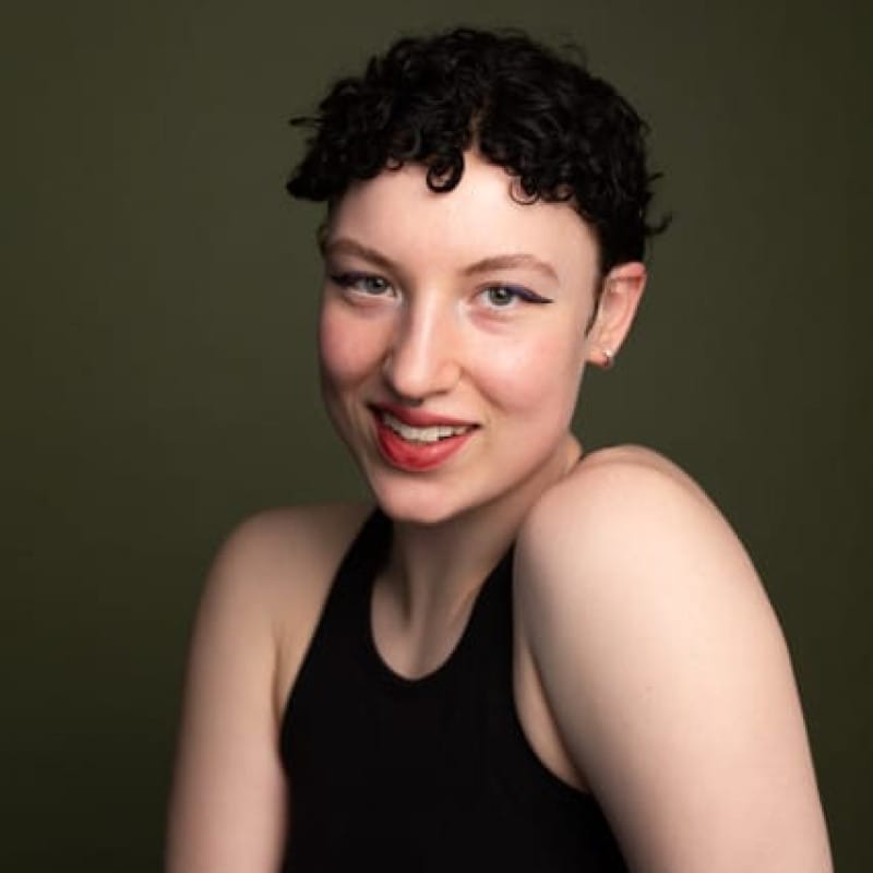 Headshot of a person with short, curly hair, warmly smiling at the camera against a dark green background.