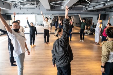 A large group of participants in a dance workshop stand in a circle, with one arm raised above their heads.