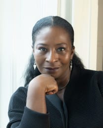 Headshot of Sharon Watson, Candoco's new Chair, smiling gently while resting her chin on her hand. She wears a black jumper and silver earrings.