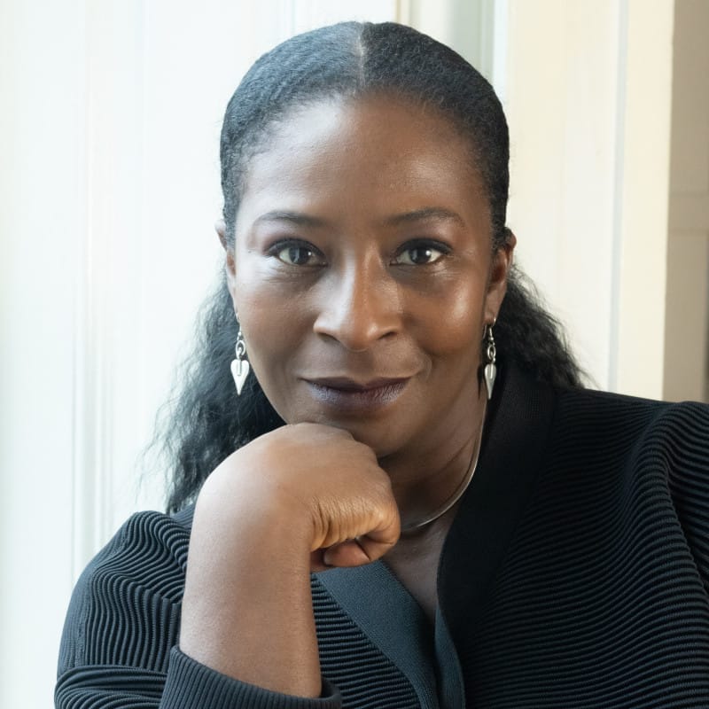 Headshot of Sharon Watson, Candoco's new Chair, smiling gently while resting her chin on her hand. She wears a black jumper and silver earrings.