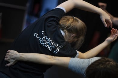 A close-up of a Candoco team member in a black t-shirt with a large white Candoco logo on the back. They are leaning to one side with their arms raised above their head and hands almost touching, while another dancer supports them with one arm around their side. The photo is taken from behind.