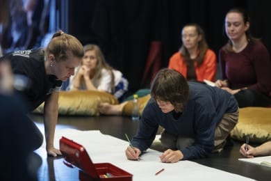 A group of participants is engaging in a workshop. The focus is on two people writing on large sheets of paper on the ground. In the background, other participants observe whilst resting on bean bags.
