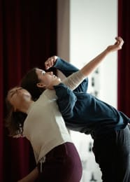 Close-up of two young people dancing indoors. One person, facing the camera, leans backward with eyes closed and one arm extended gracefully to the side. Their dance partner stands with their back against them, holding onto the extended arm for support, creating a fluid, synchronised pose.