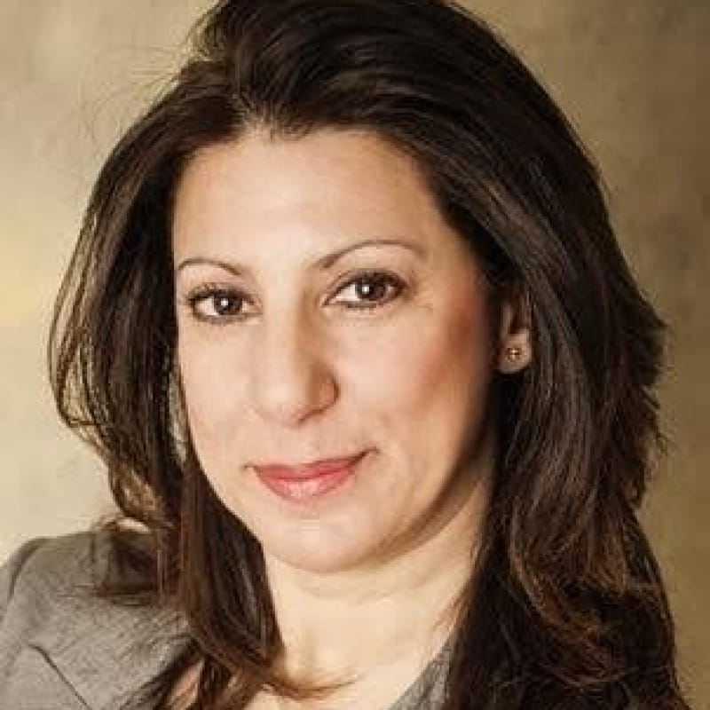 Headshot of a person with long, straight brown hair, wearing a grey jacket and a matching top, smiling gently at the camera.