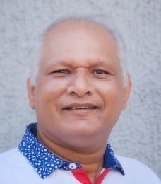 A person with short white hair, wearing a white shirt with blue and white patterns, gently smiling at the camera.