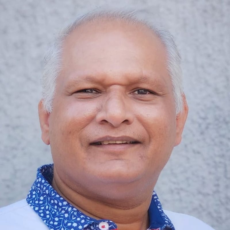 A person with short white hair, wearing a white shirt with blue and white patterns, gently smiling at the camera.