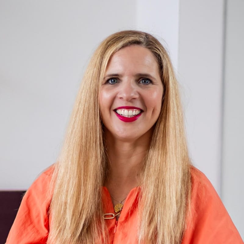 Headshot of a person with long blonde hair, leaning against a wooden banister while wearing a salmon-orange dress with wide sleeves.