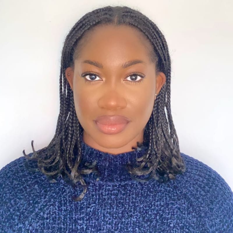 Headshot of a person with short braided hair wearing a blue jumper, looking directly at the camera with a serious expression.