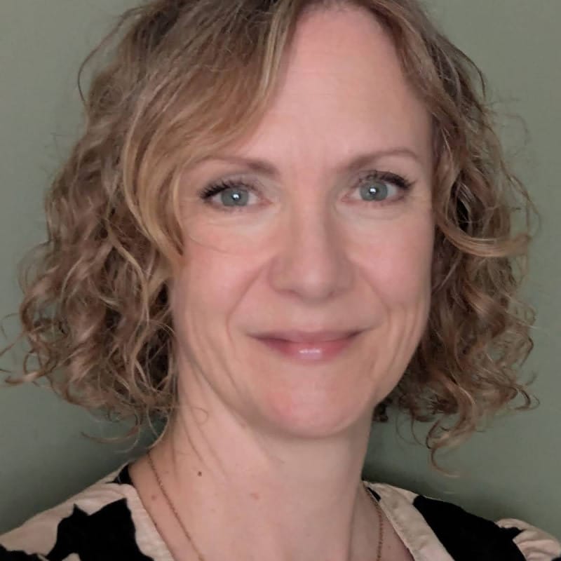 A softly lit headshot of a person with short, blonde curly hair, giving a gentle smile and gazing straight into the camera, with natural tones and a clean pale green background.