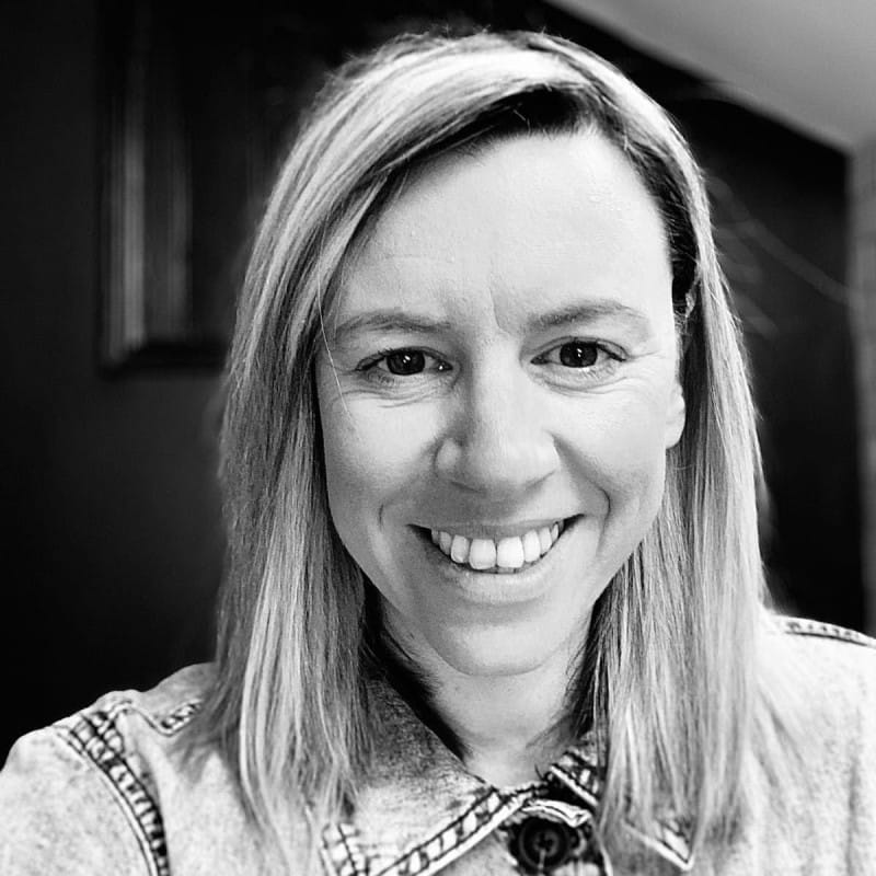 Black and white headshot of a person with mid-length straight hair smiling warmly at the camera against a dark background.