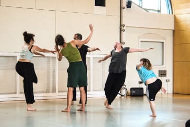 Five dancers move dynamically in a bright, spacious studio, some leaning forward and others backward with their arms raised mid-motion.