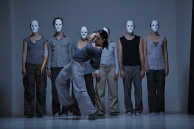 Six dancers stand in a line wearing blank white masks. Another performer without a mask bends backwards in front of them