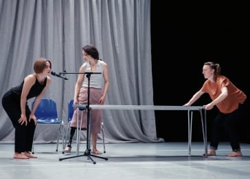 Three performers are on a stage with a grey draped back cloth. There is a table in the middle and one dance is pushing down on the table. Another leans toward a microphone while the last dancer watches on.
