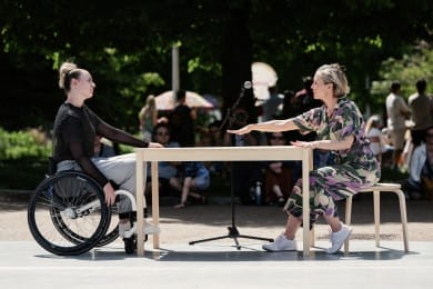 Two dancers are performing outside with trees behind them and a table in between. One dancer is reaching across the table to the other.