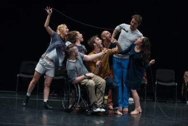 A group of performers gathers around a microphone on a dark stage, leaning in and speaking with animated expressions. One performer uses a wheelchair, and the group shares a playful, energetic sense of connection.