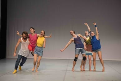 Six dancers in colourful costumes are moving on a bare stage.