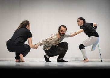 Three dancers crouch on a stage with a white backdrop. Two dancers hold the hands of the dancer in the centre.