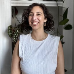 A person with short curly hair smiles warmly at the camera, standing indoors in front of shelves with plants.