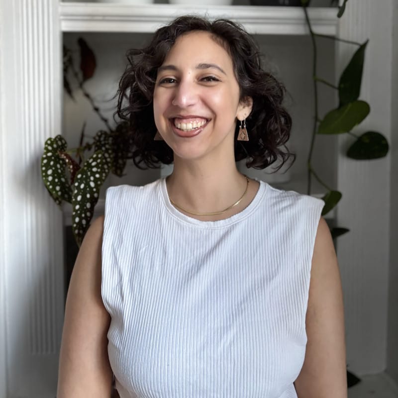 A person with short curly hair smiles warmly at the camera, standing indoors in front of shelves with plants.