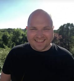 A headshot of a smiling Tom Curteis taken outside with green trees and a blue sky behind him.