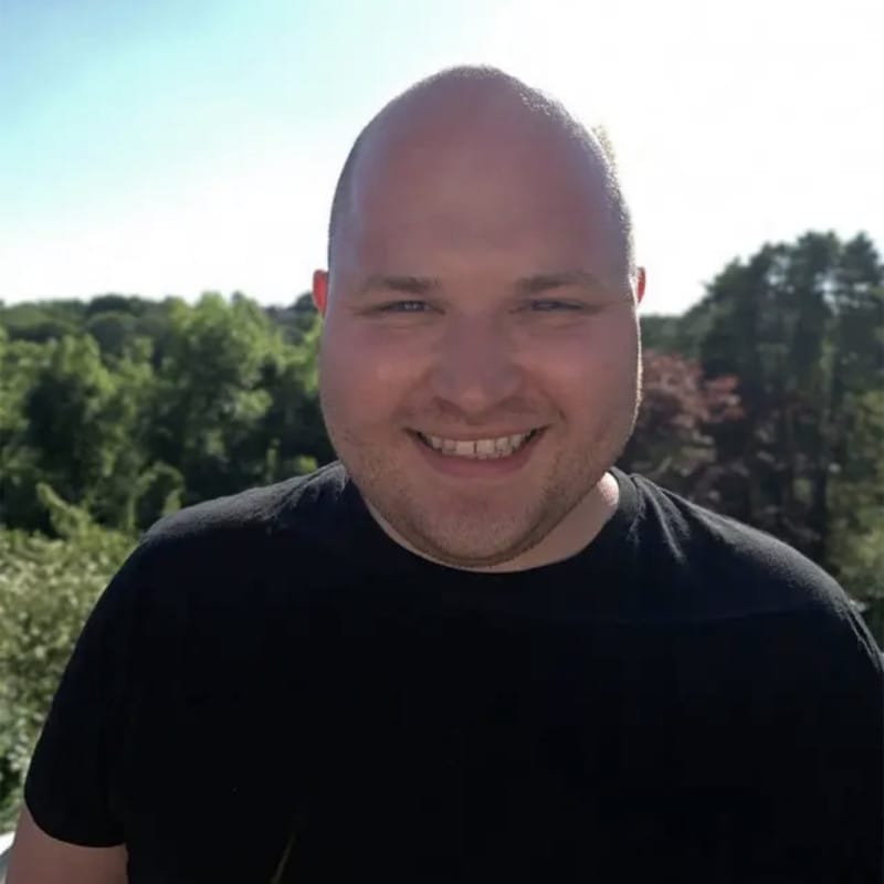A headshot of a smiling Tom Curteis taken outside with green trees and a blue sky behind him.